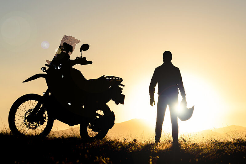 A man stands next to his motorcycle while enjoying the sunset over the mountains.