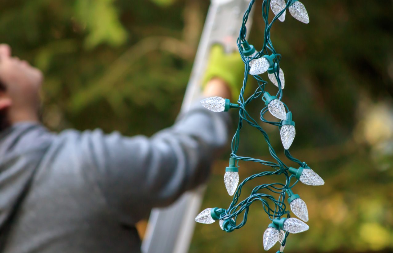 Homeowner in the process of scaling a ladder while holding a bundle of Christmas lights.