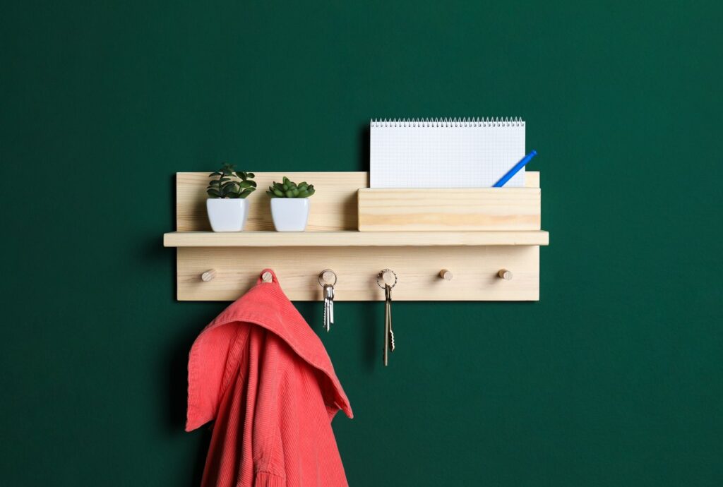 A light colored wooden key hook hangs on a dark green wall. The hooks hold two sets of keys and a pink jacket. A notebook and two small plants in white pots wit on the self.