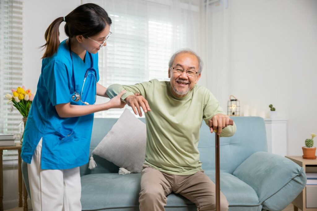 A young nurse helps an older man with a cane off a couch.