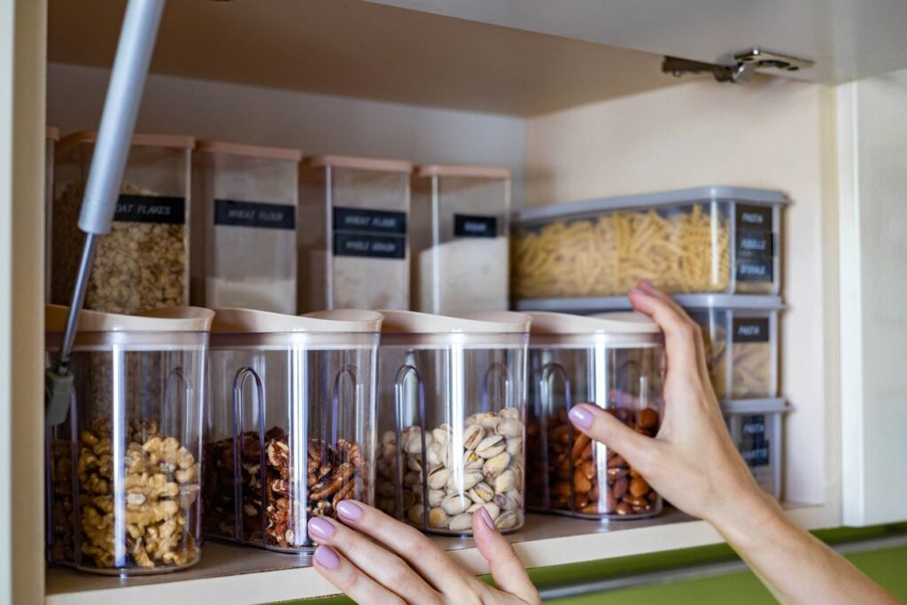 A pair of hands reaches for organized bins of assorted nuts in a kitchen cabinet.