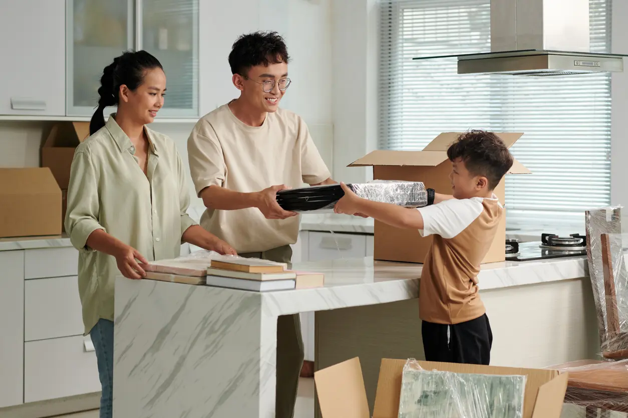 A young family places framed photos and books into cardboard boxes while in their home kitchen.