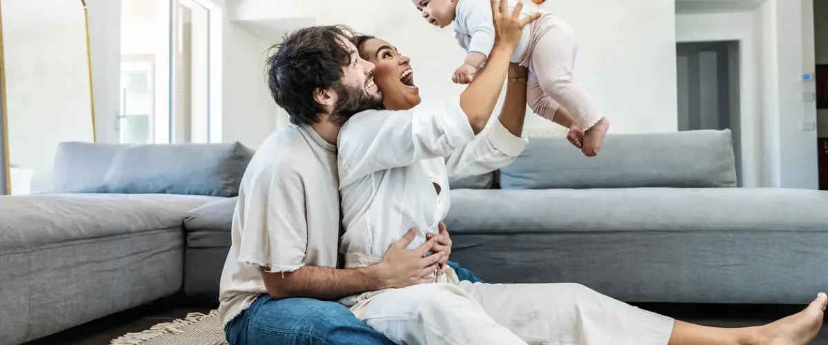 A young couple plays with their baby as they sit on the floor in their living room.