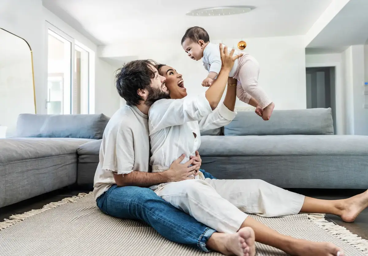 A young couple plays with their baby as they sit on the floor in their living room.