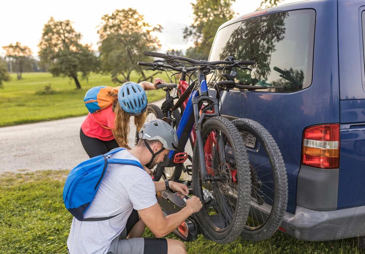 Two people in bike helmets are taking mountain bikes off the vehicle rack of a parked blue van on a sunny day.