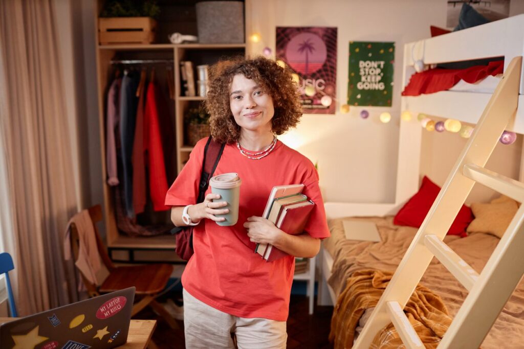 A young woman attending college stands in her neat dorm room, books and coffee in hand.