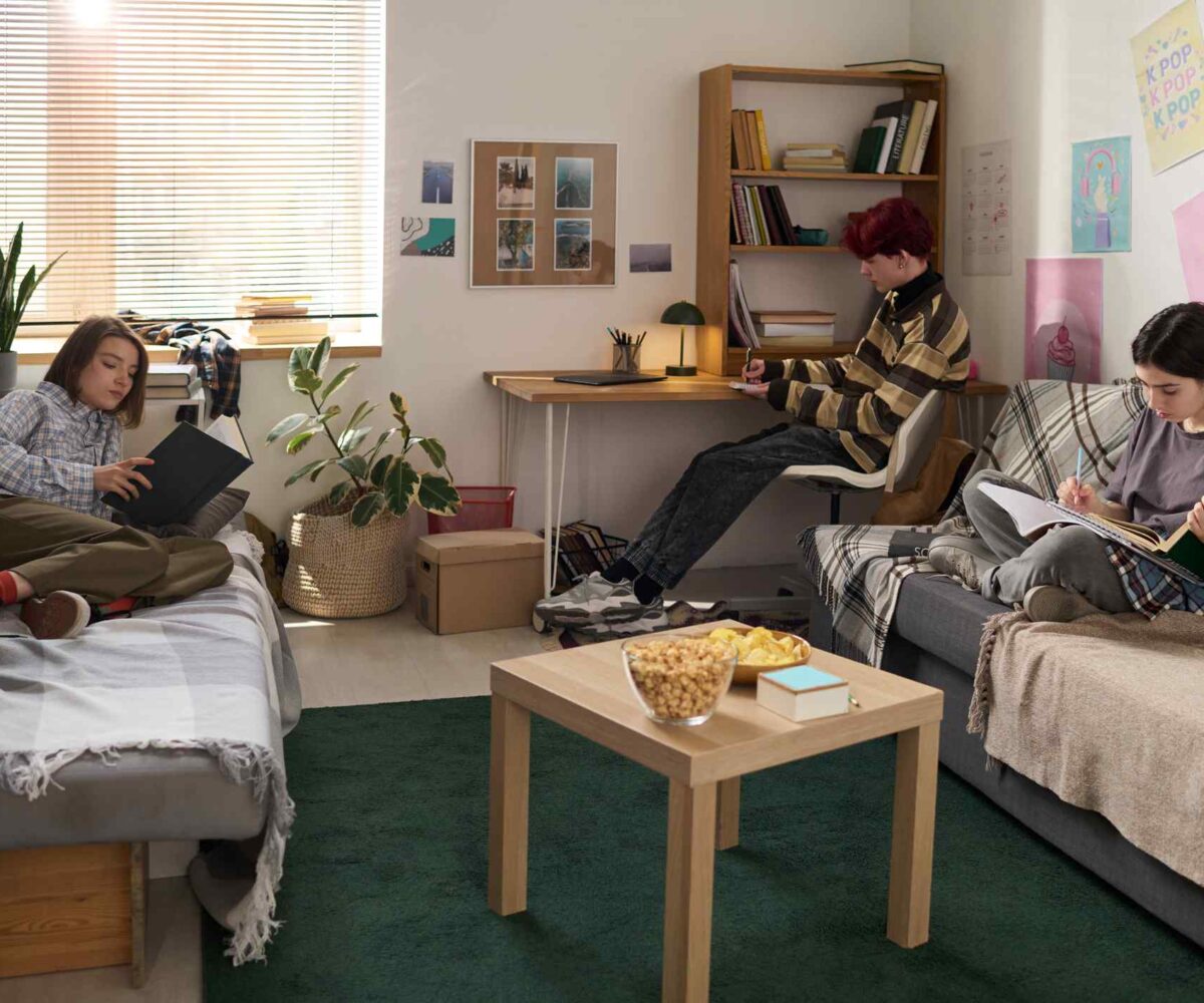 Three college students sit, reading and studying, inside a dorm room.