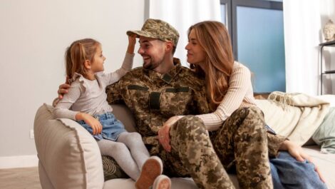 A military member in uniform sitting on the couch with his daughter and wife.